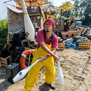 A person in a red hat and yellow overalls juggling pins in front of a quirky, nautical-themed backdrop